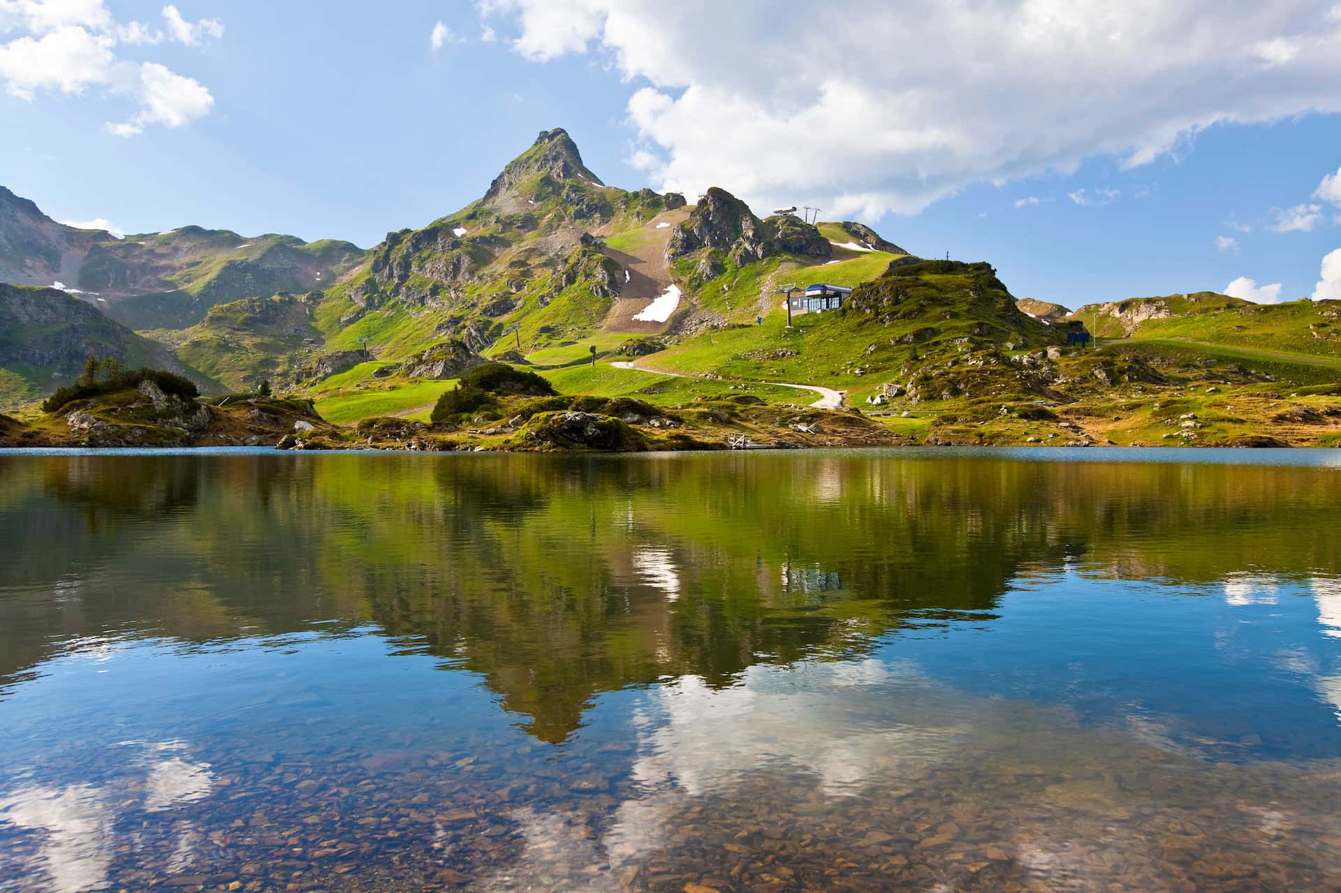 Alpine lake reflecting green mountains under a blue sky with clouds; hiking trail visible.