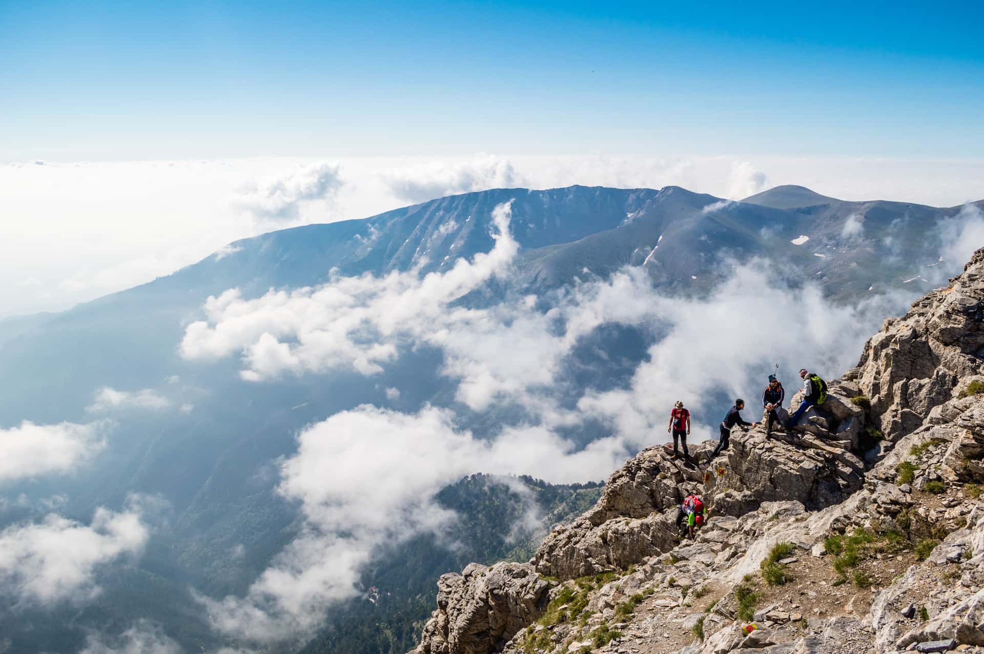 Hikers resting on rocky mountain ridge above clouds with distant peaks under bright blue sky