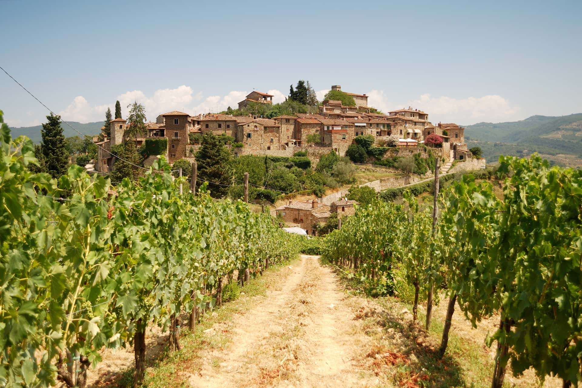 Vineyard path leading to historic stone village on hillside under blue sky in Tuscany.