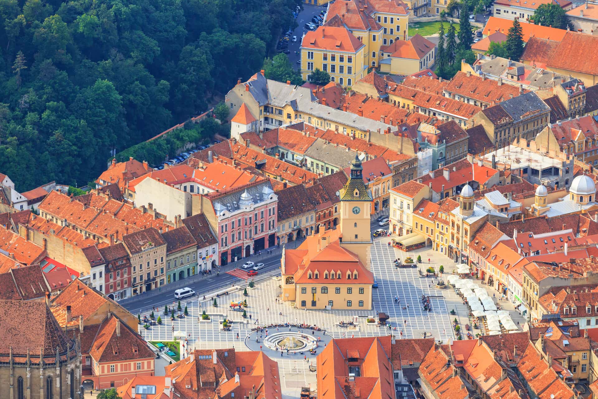 Aerial view of Brasov's Council Square with clock tower, red roofs, and surrounding green forest.