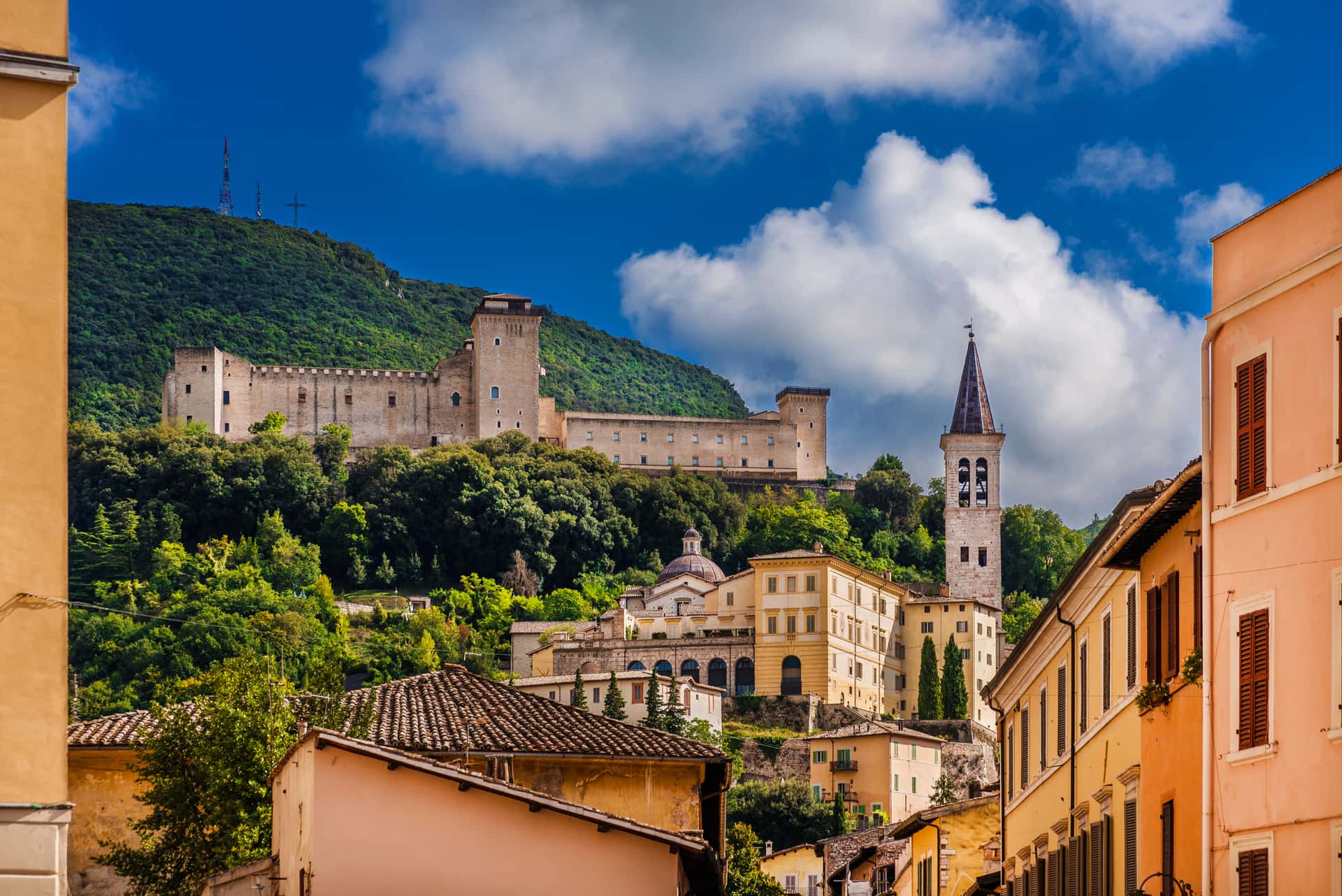 Fortress on a green hill above historic town buildings with a prominent bell tower.