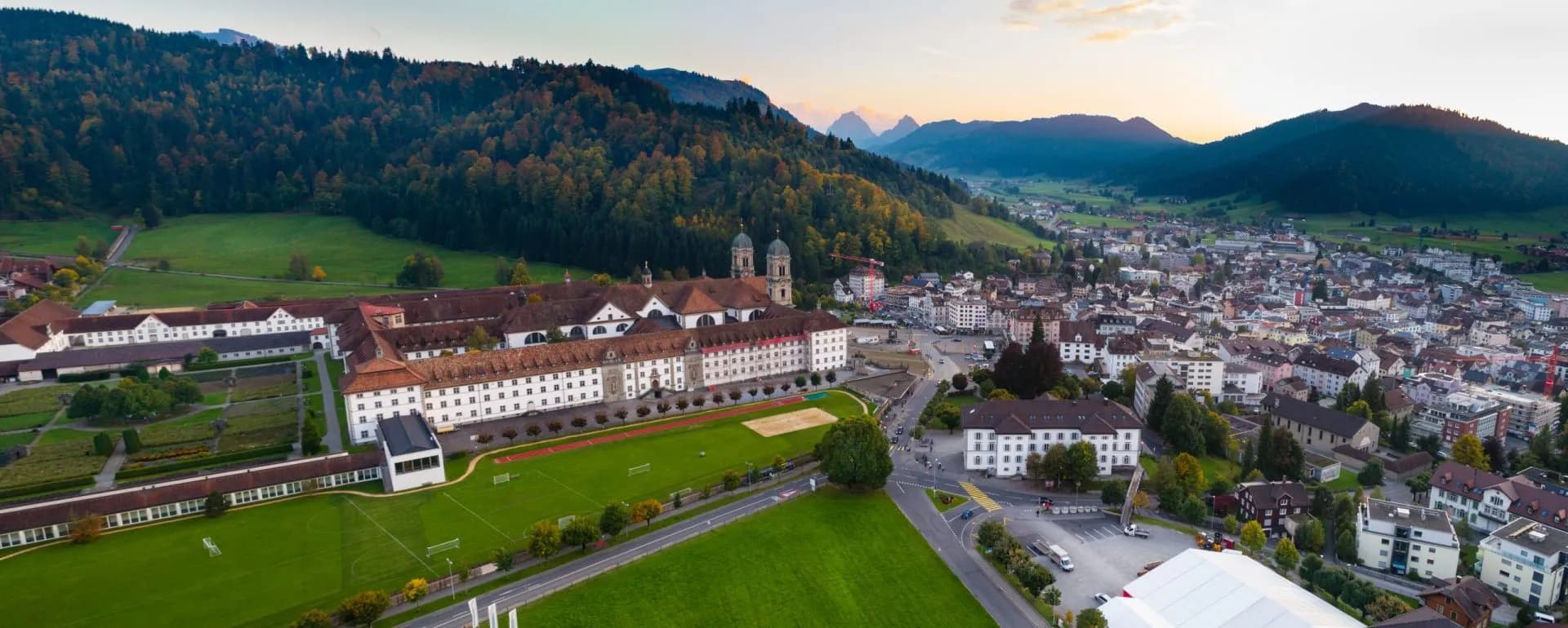 Aerial view of Einsiedeln Abbey and town nestled in a valley with forested mountains at sunset.