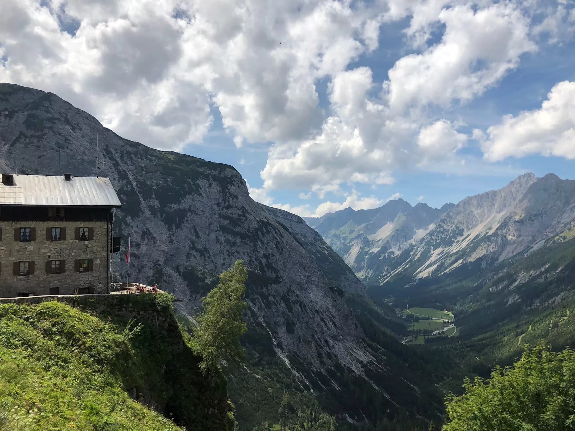 Stone mountain hut perched on a cliff overlooking an alpine valley and distant peaks.