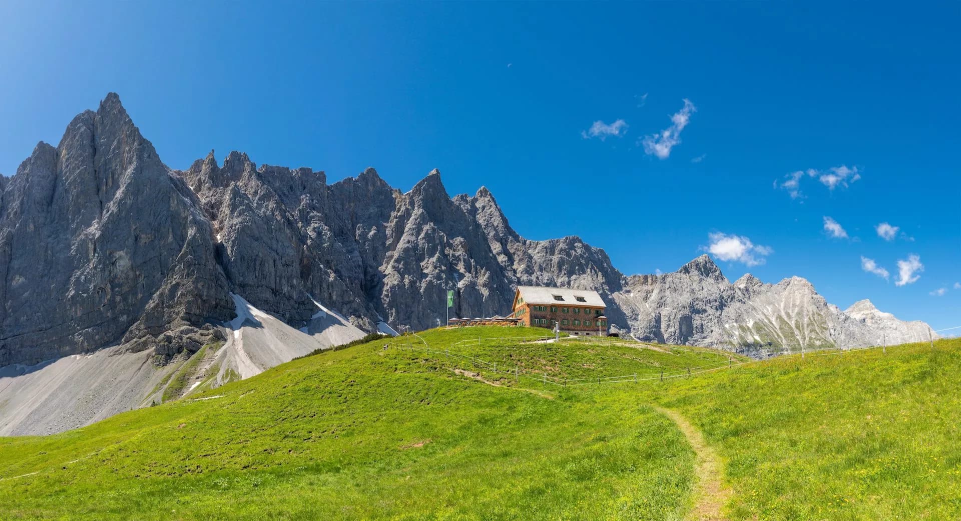 Mountain hut on grassy alpine meadow below jagged gray peaks under bright blue sky