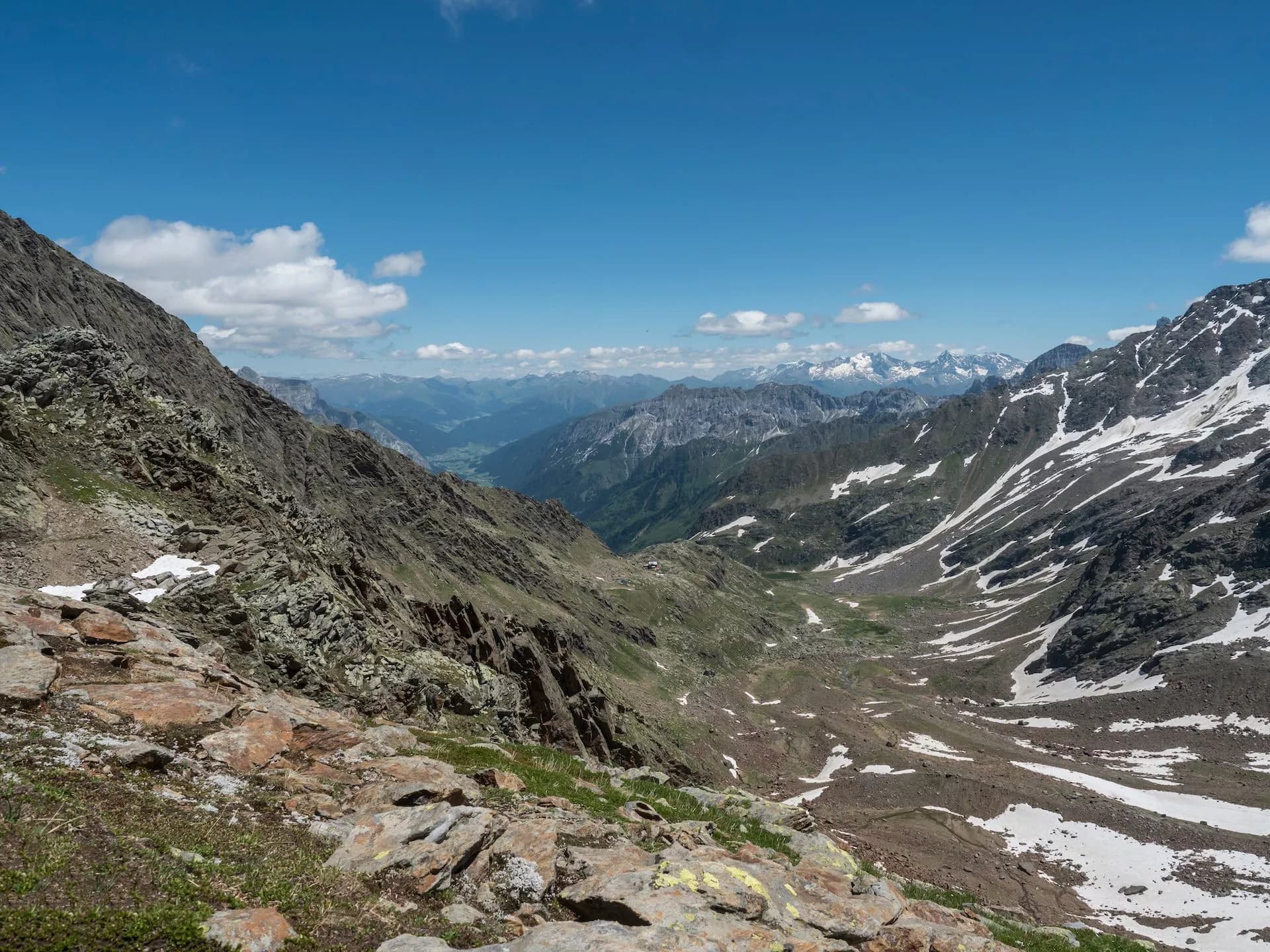 Rocky alpine landscape with patches of snow, green valley below, and distant snow-capped mountains under blue sky.