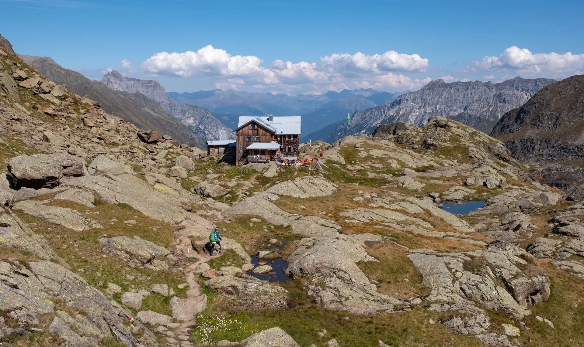 Wooden mountain hut near rocky terrain with a hiker approaching on a sunny day, Bremer Hütte.