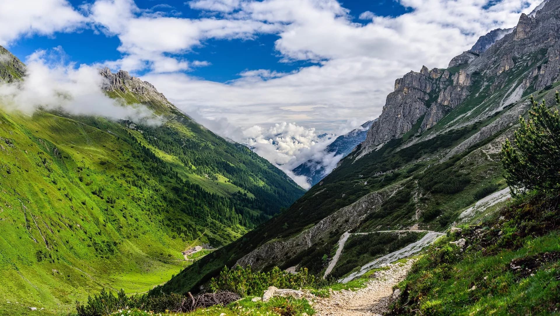 Hiking trail through steep green valley between rocky mountains under cloudy blue sky in Pinnistal Valley.