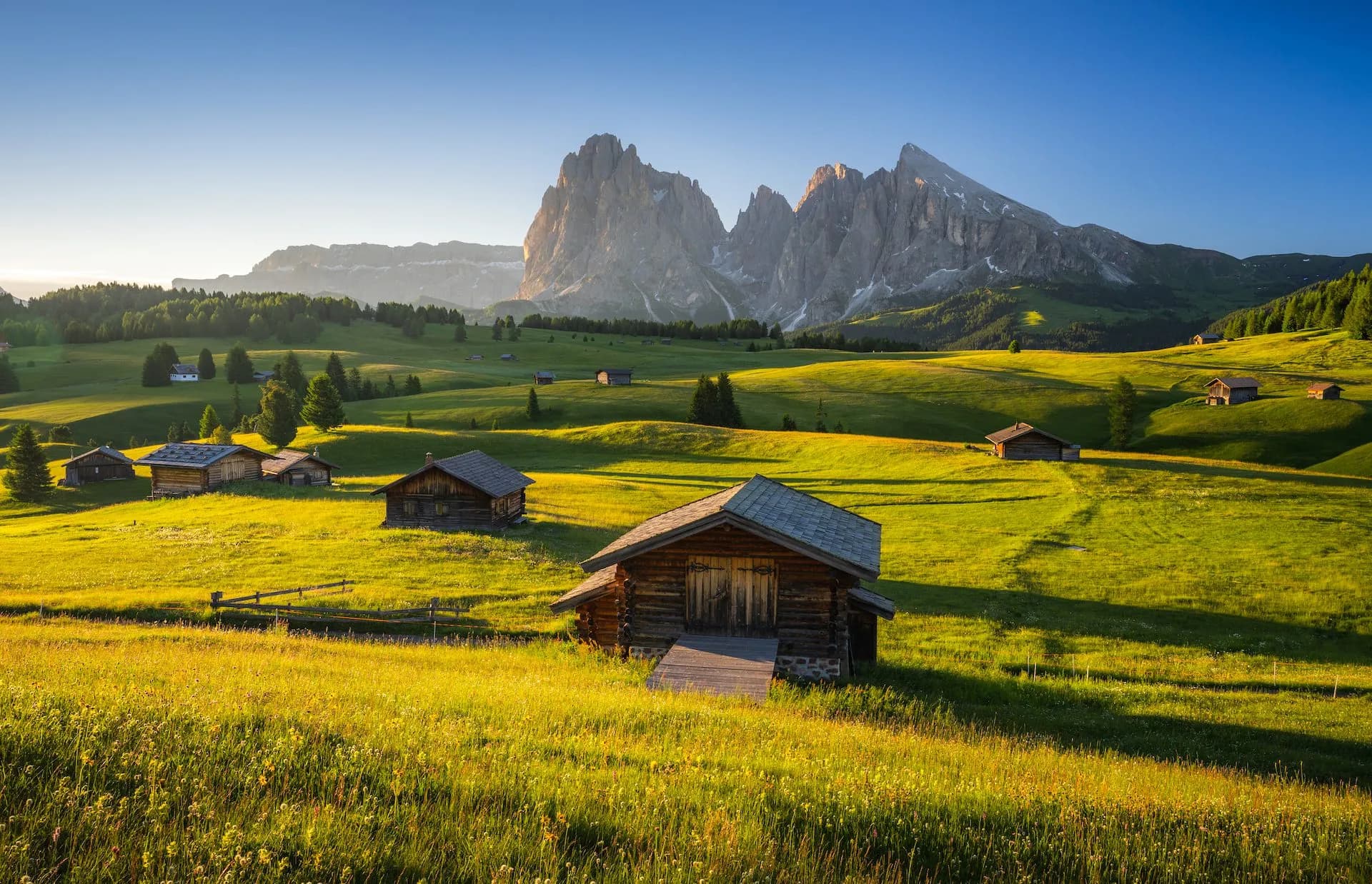 Wooden alpine huts on sunny green meadows below rugged mountain peaks at Seiser Alm.