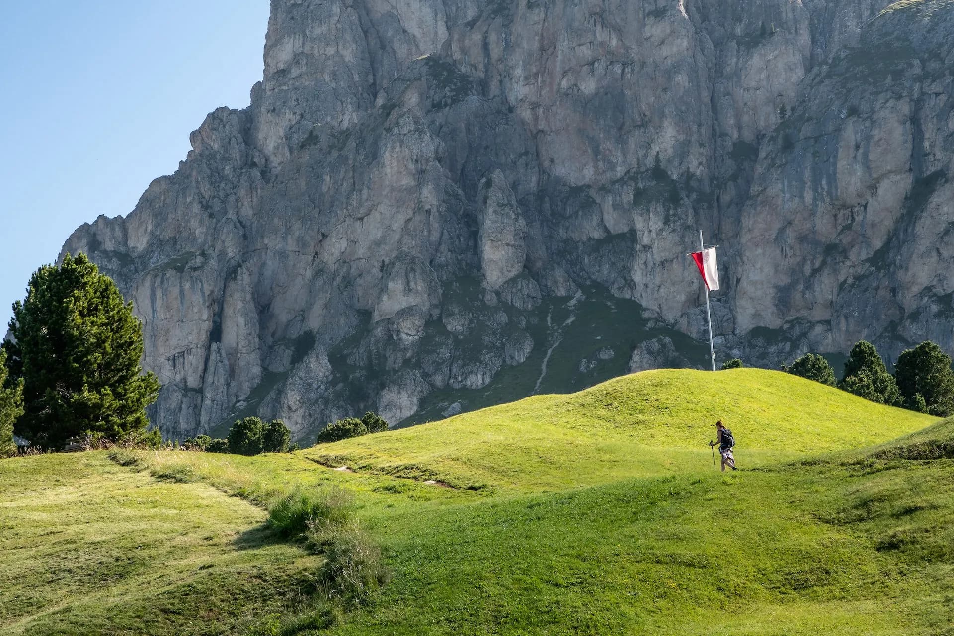 Hiker with poles on grassy hill below massive rock face with red and white flag near Saltria.