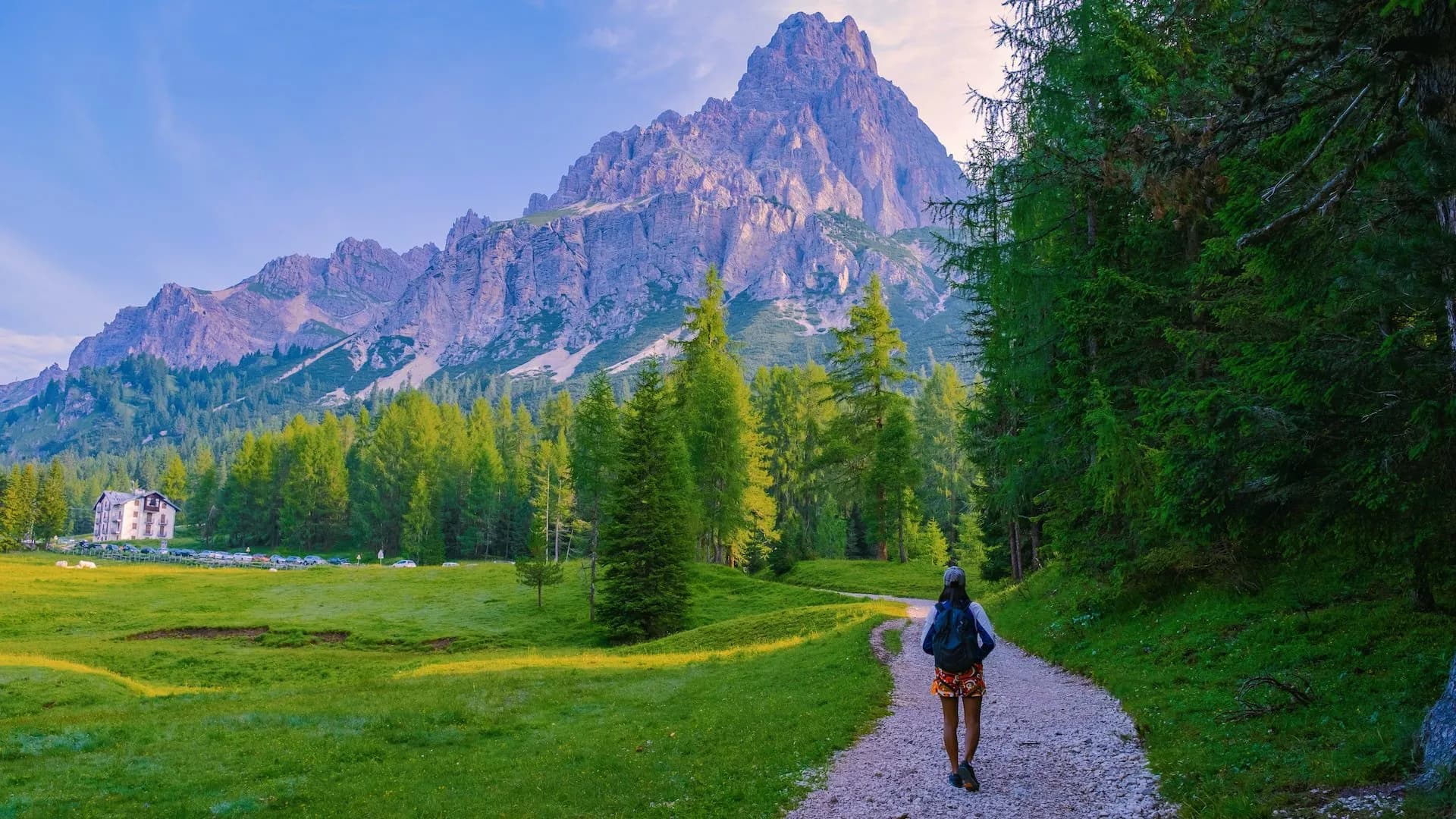 Hiker walking on gravel path toward massive Dolomite mountain peak and green forest.