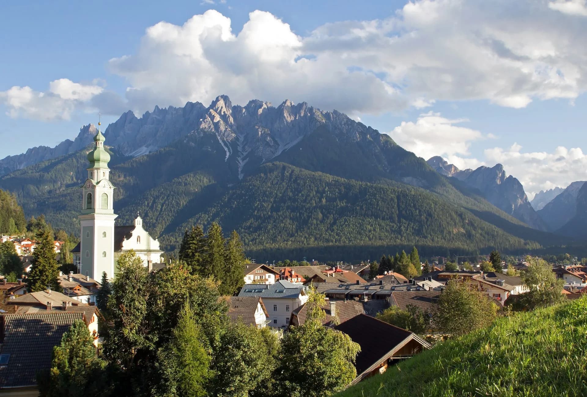 Church tower overlooking Dobbiaco village with forested mountains under cloudy sky.
