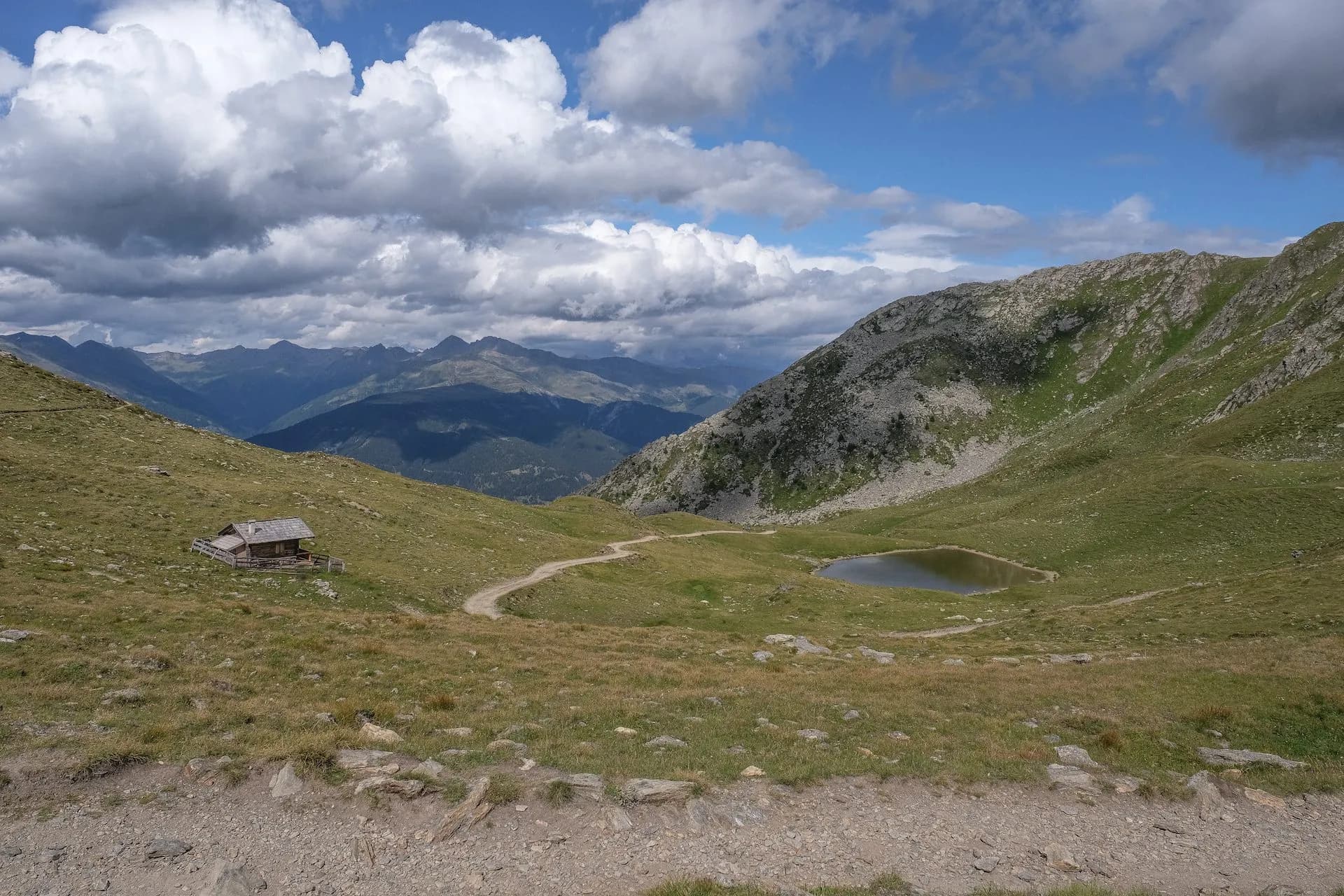 Alpine hiking trail past a small hut toward a mountain lake under dramatic clouds