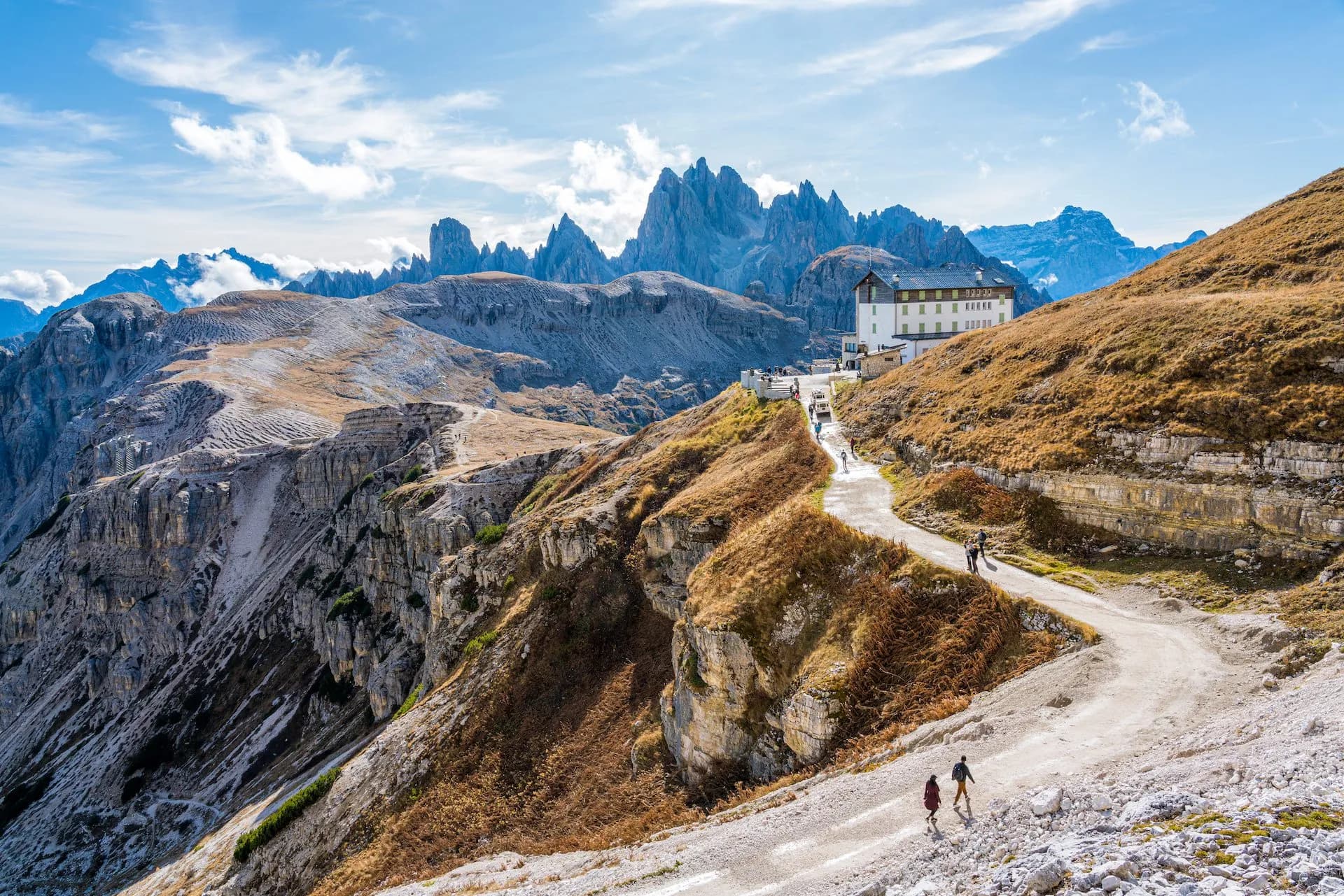 Rifugio Auronzo mountain lodge on a winding path with hikers, set against jagged Dolomite peaks.