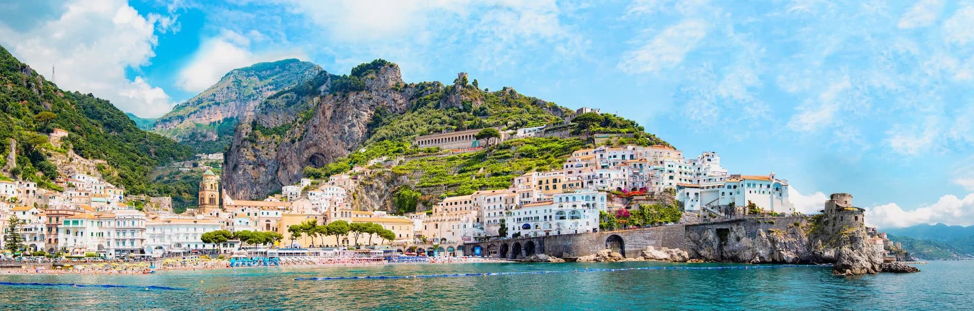 Coastal town with white buildings climbing steep green mountains above a crowded beach and turquoise sea.