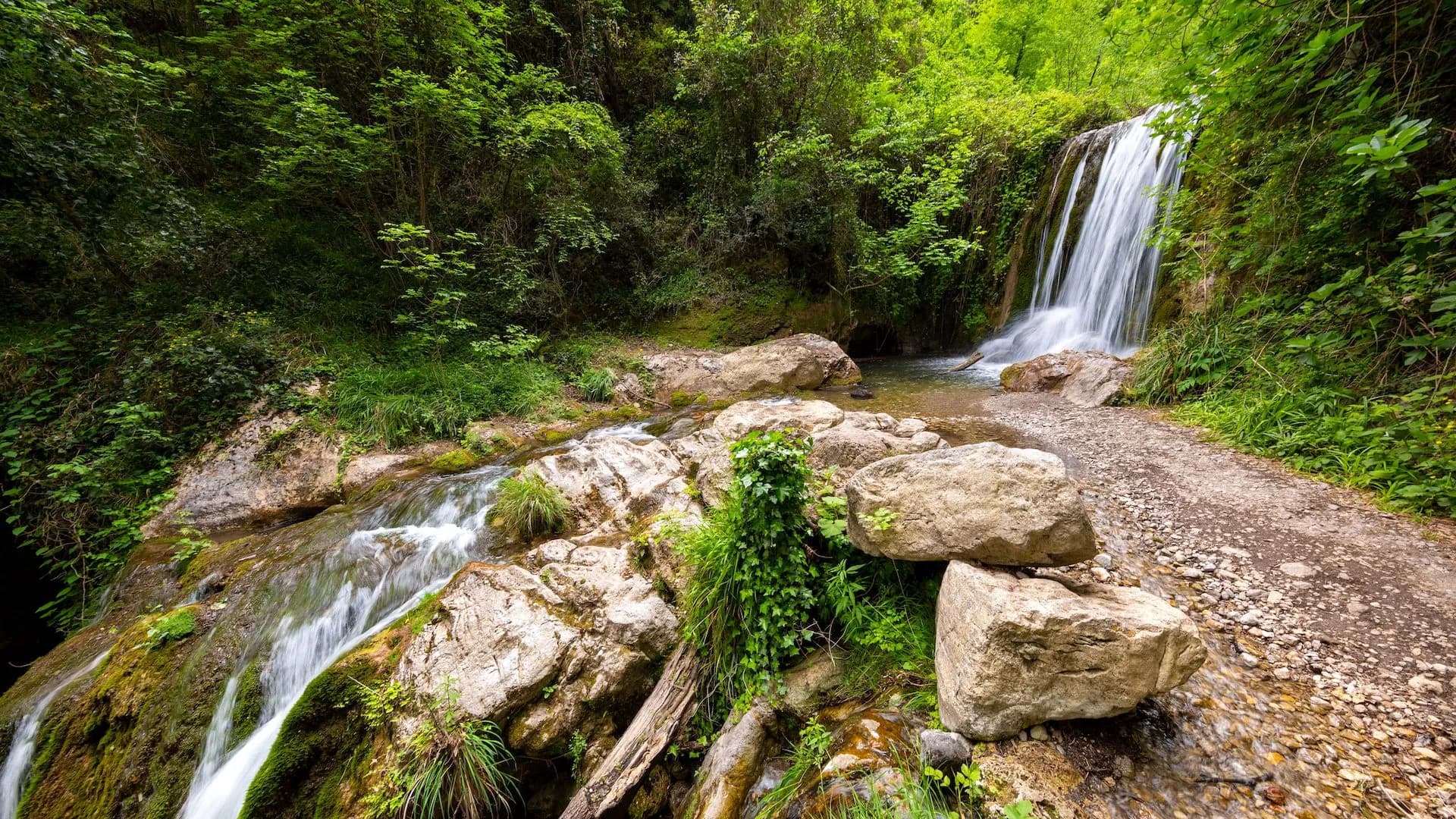 Waterfall cascading into a stream with mossy rocks and a dirt path in the Valley of the Ferriere.