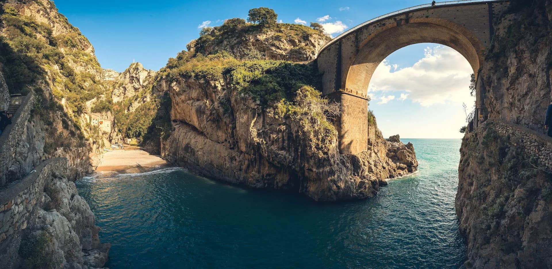 Stone arch bridge over turquoise sea inlet with steep cliffs and a small beach.