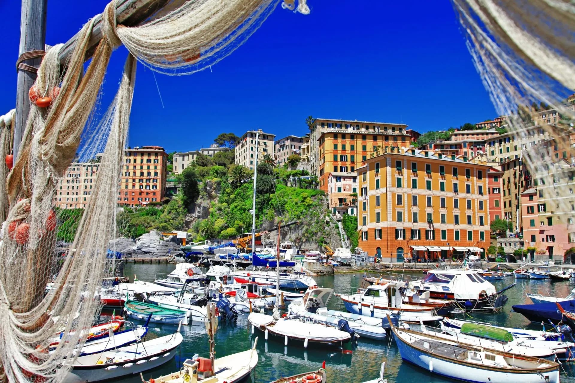 Fishing nets frame boats docked in the harbor below colorful buildings in Camogli, Liguria.