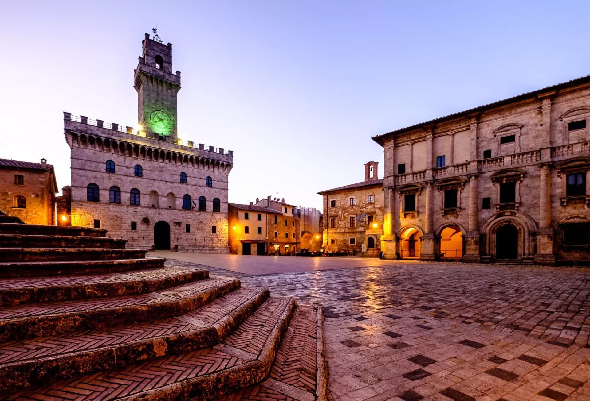 Historic town square with illuminated clock tower and stone steps at dusk in Montepulciano.