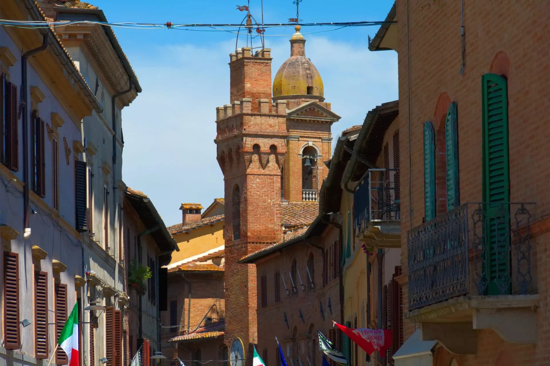 Brick bell tower visible between buildings on a narrow street in Buonconvento, Italy.