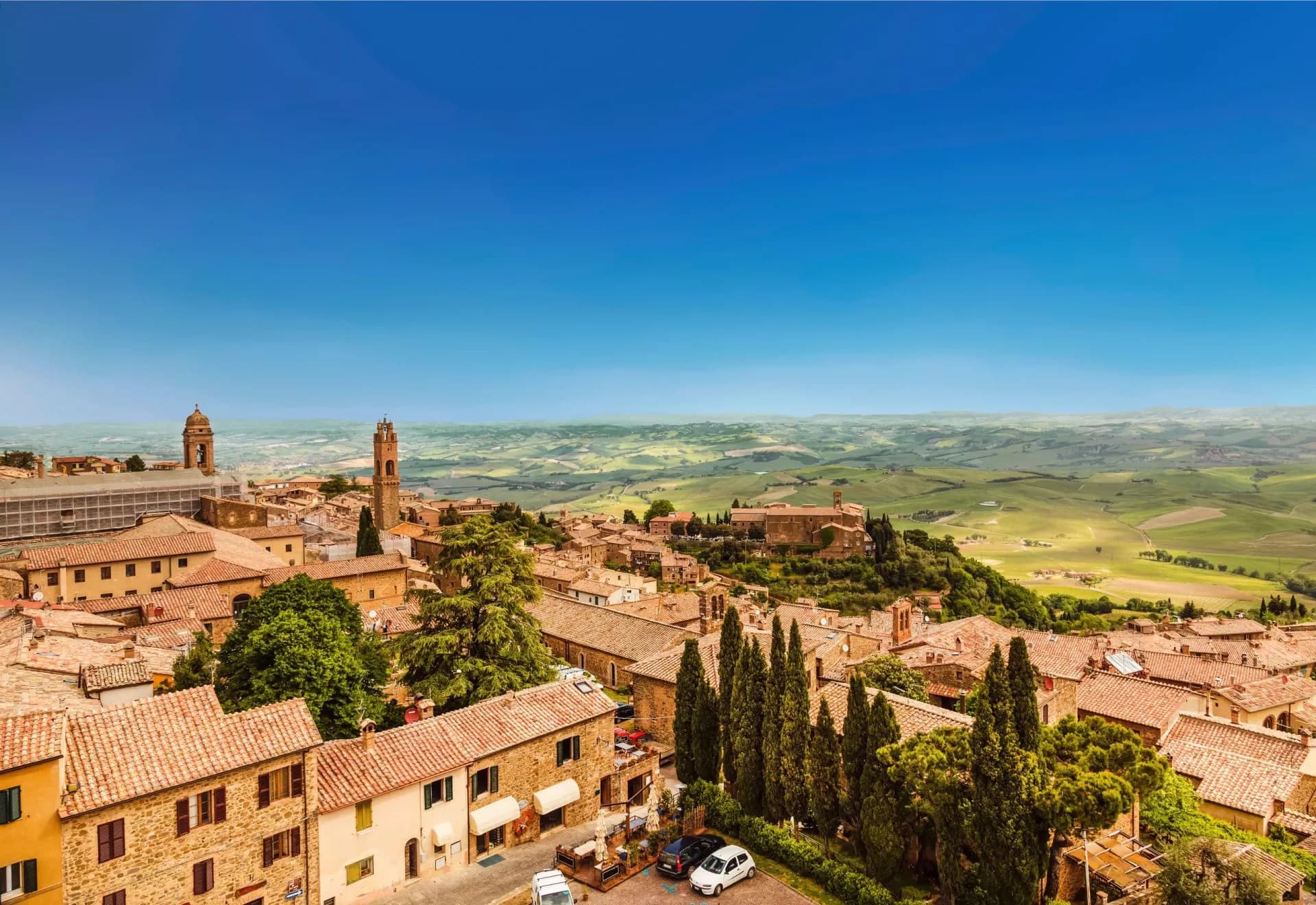 Montalcino hill town with terracotta roofs overlooking rolling green Tuscan countryside under a clear blue sky.