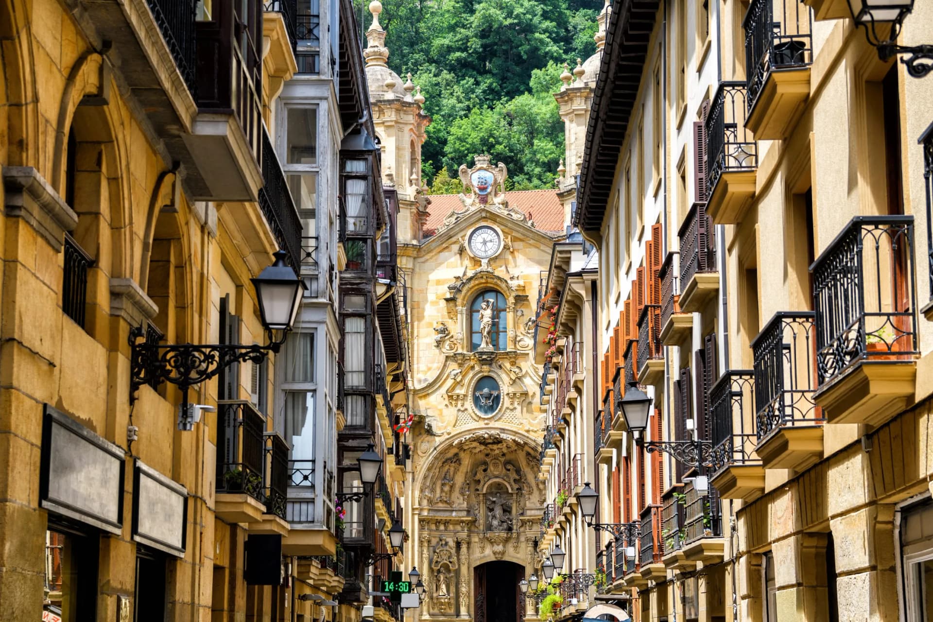 Narrow street view of ornate church facade in San Sebastian Old Town with green hillside background.