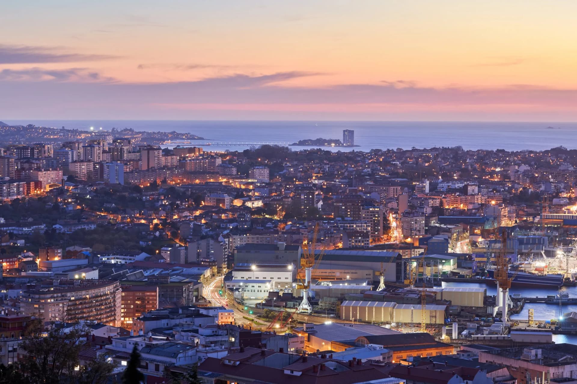 City of Vigo at sunset with illuminated buildings, shipyard cranes, and the sea in the background.