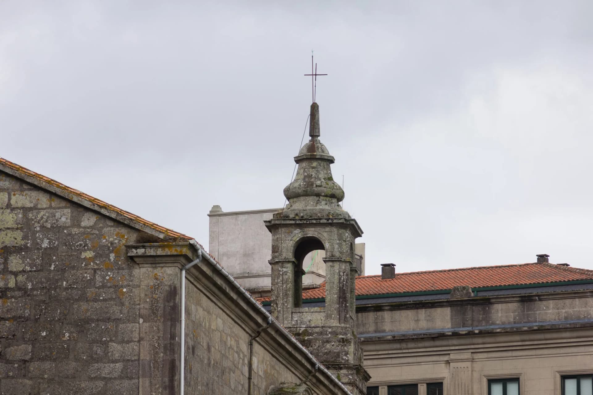 Stone bell tower with a cross atop an old building under a gray sky in Padrón.
