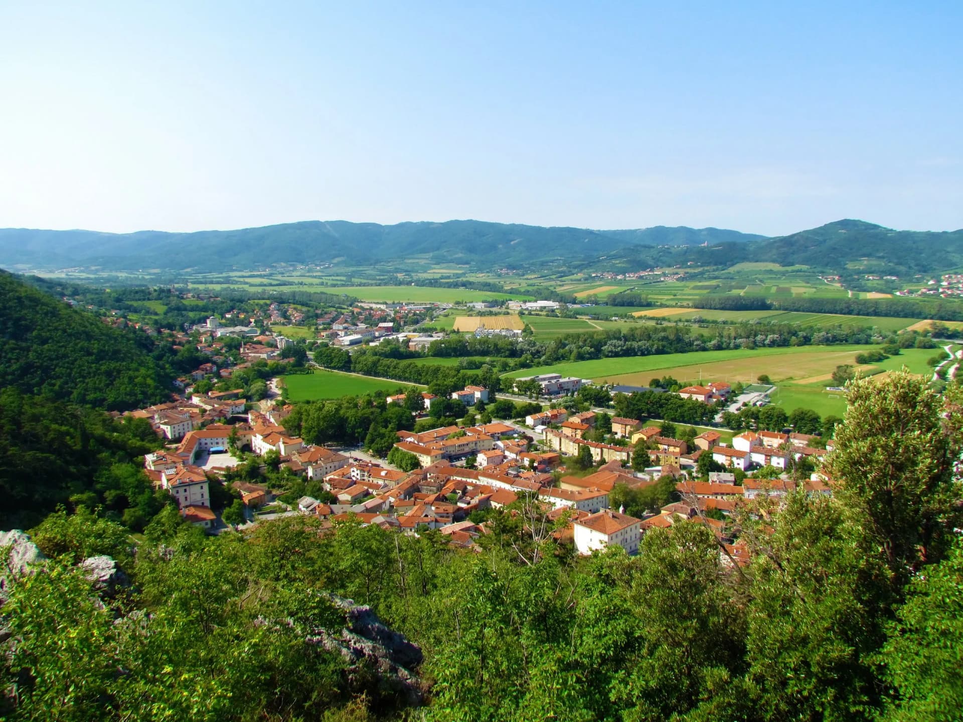 View of the small town Vipava nestled in a valley with green fields and rolling hills under a clear sky.