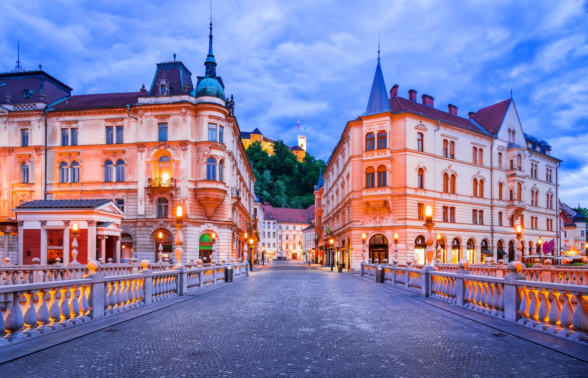 Cobblestone bridge leading toward Ljubljana Castle illuminated at dusk with ornate buildings.