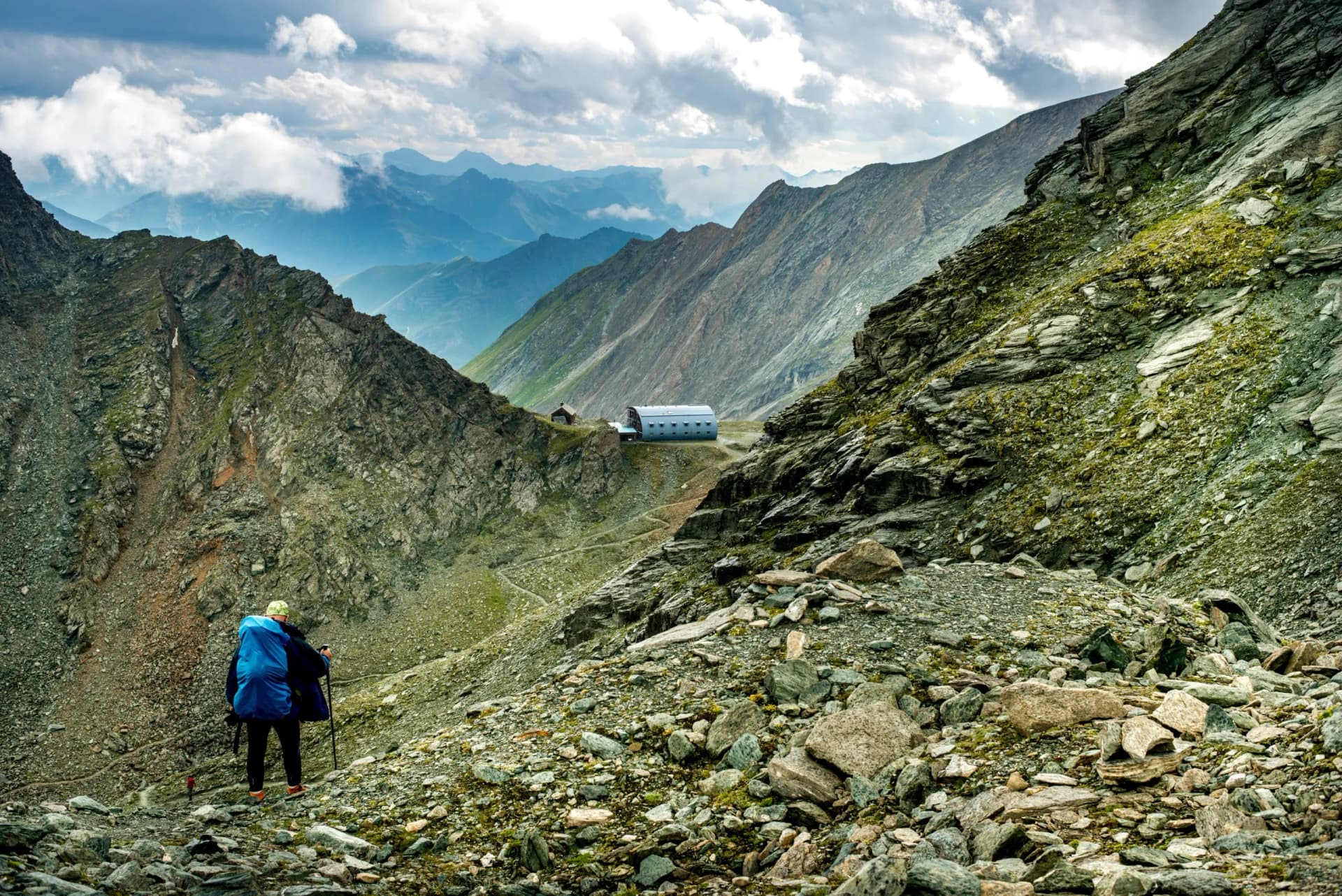 Hiker with blue backpack on rocky alpine trail approaching a mountain hut under cloudy skies.