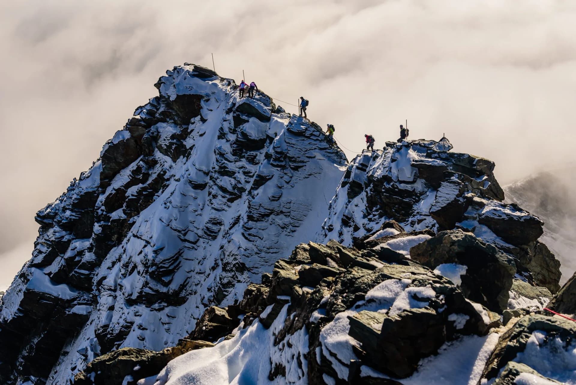 Mountaineers climbing a snowy, rocky ridge on Grossglockner peak with clouds below.