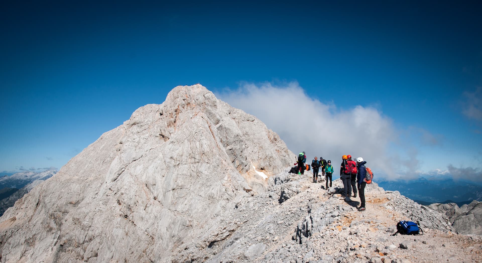Hikers with gear on rocky Triglav ridge below a massive limestone peak under a clear blue sky.