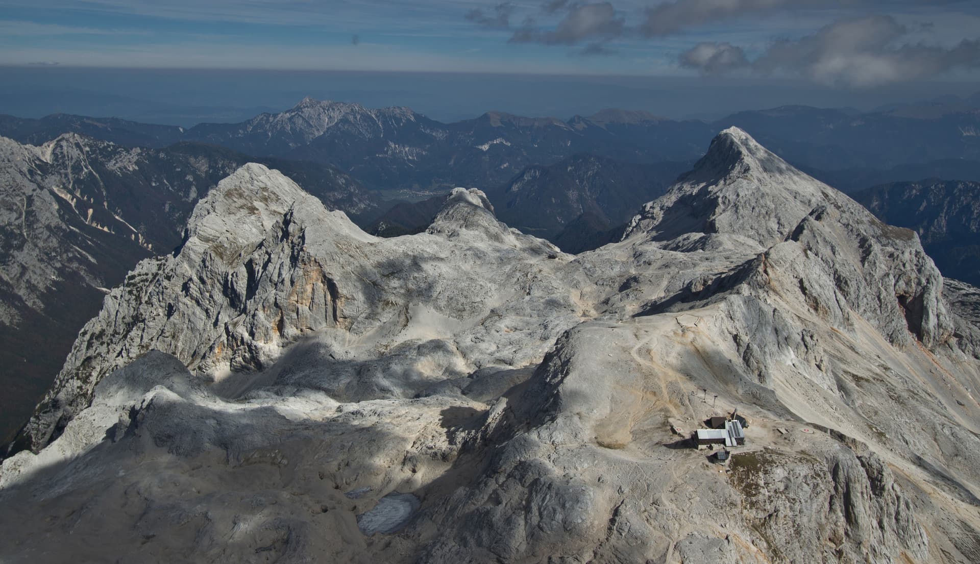 View from Triglav mountain summit showing rocky peaks, a small mountain hut, and distant ranges.
