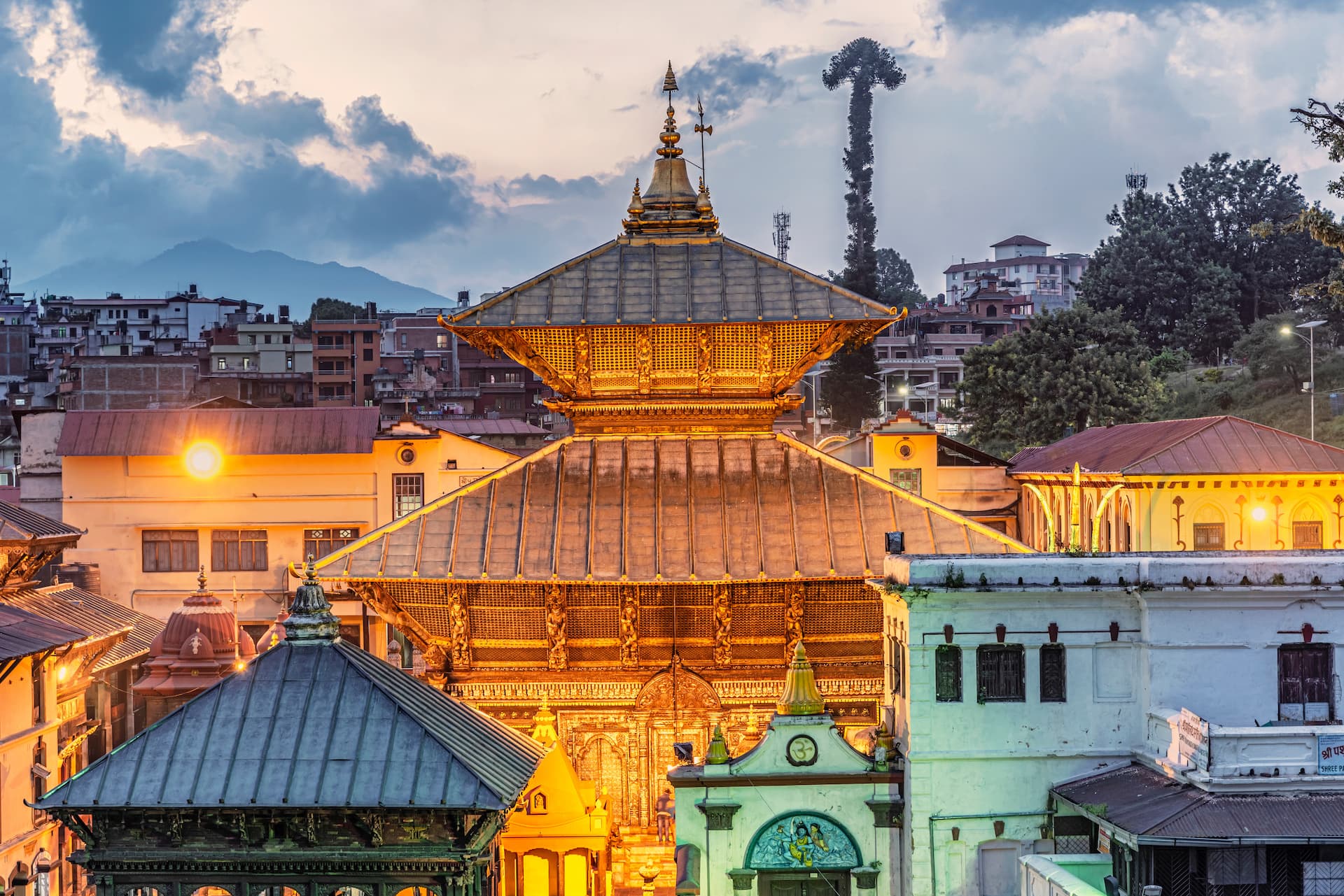 Illuminated Pashupatinath Temple roof structure against city buildings and mountains at dusk.