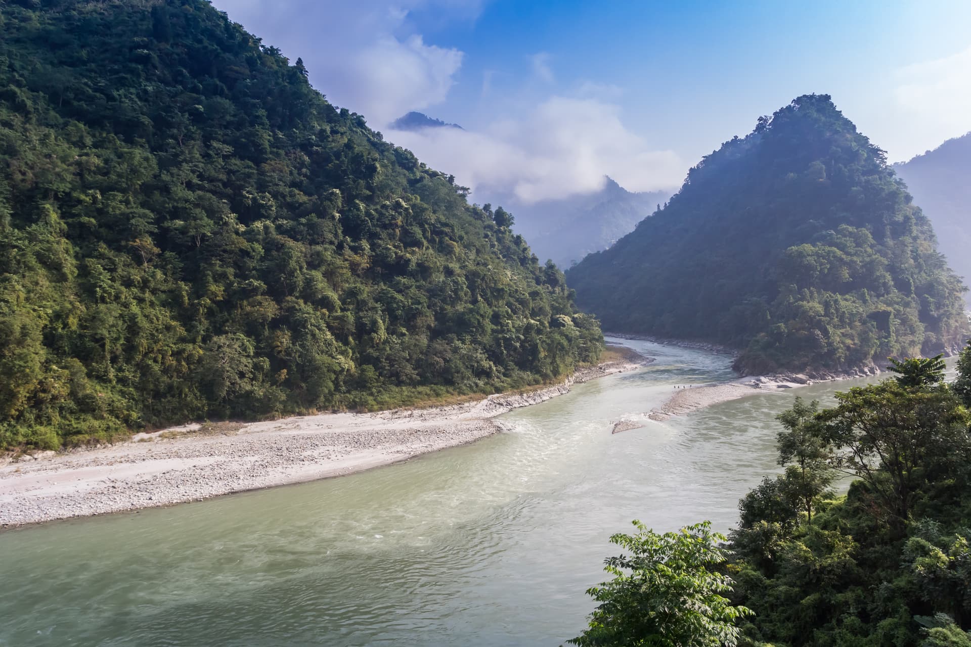 Trishuli River winding through steep, densely forested mountains under a partly cloudy blue sky.