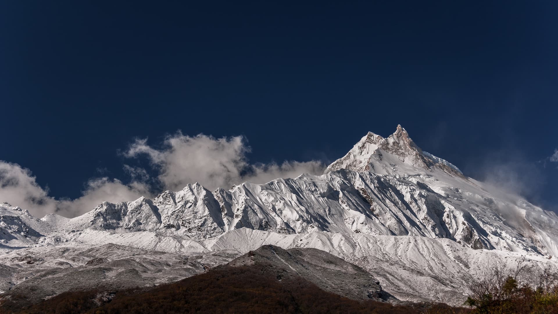 Snow-covered Manaslu mountain peaks rise above dark foothills under a deep blue sky near Samagaon.