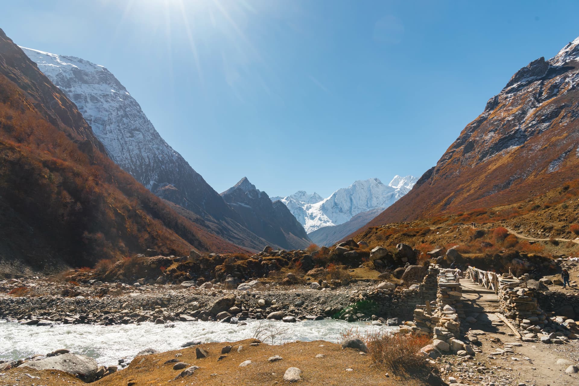 Stone bridge over rushing river in Manaslu valley with snow-capped mountains under bright sun.