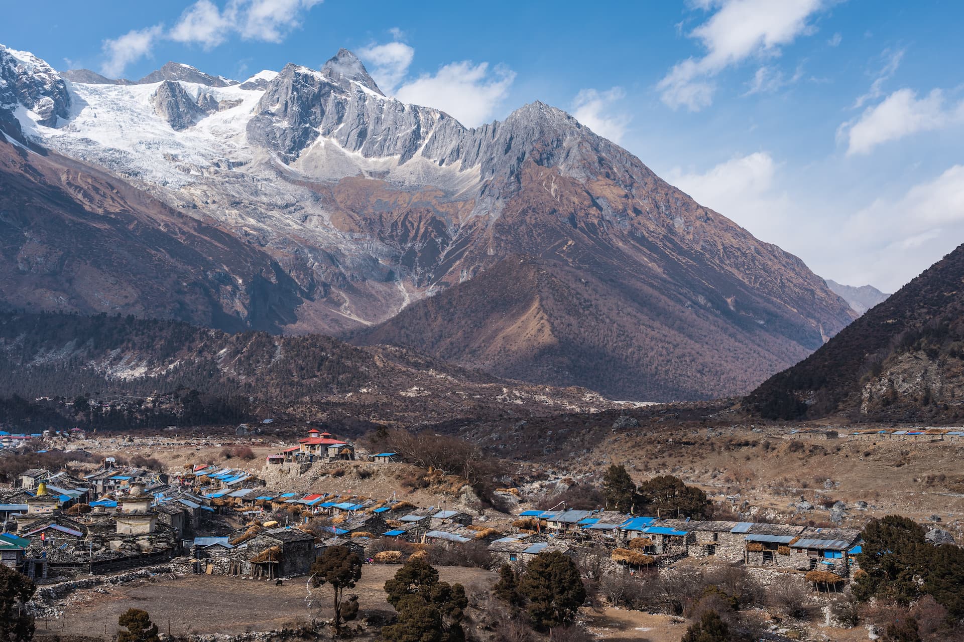 Mountain village of Samagaon nestled below massive snow-capped Himalayan peaks under a blue sky.