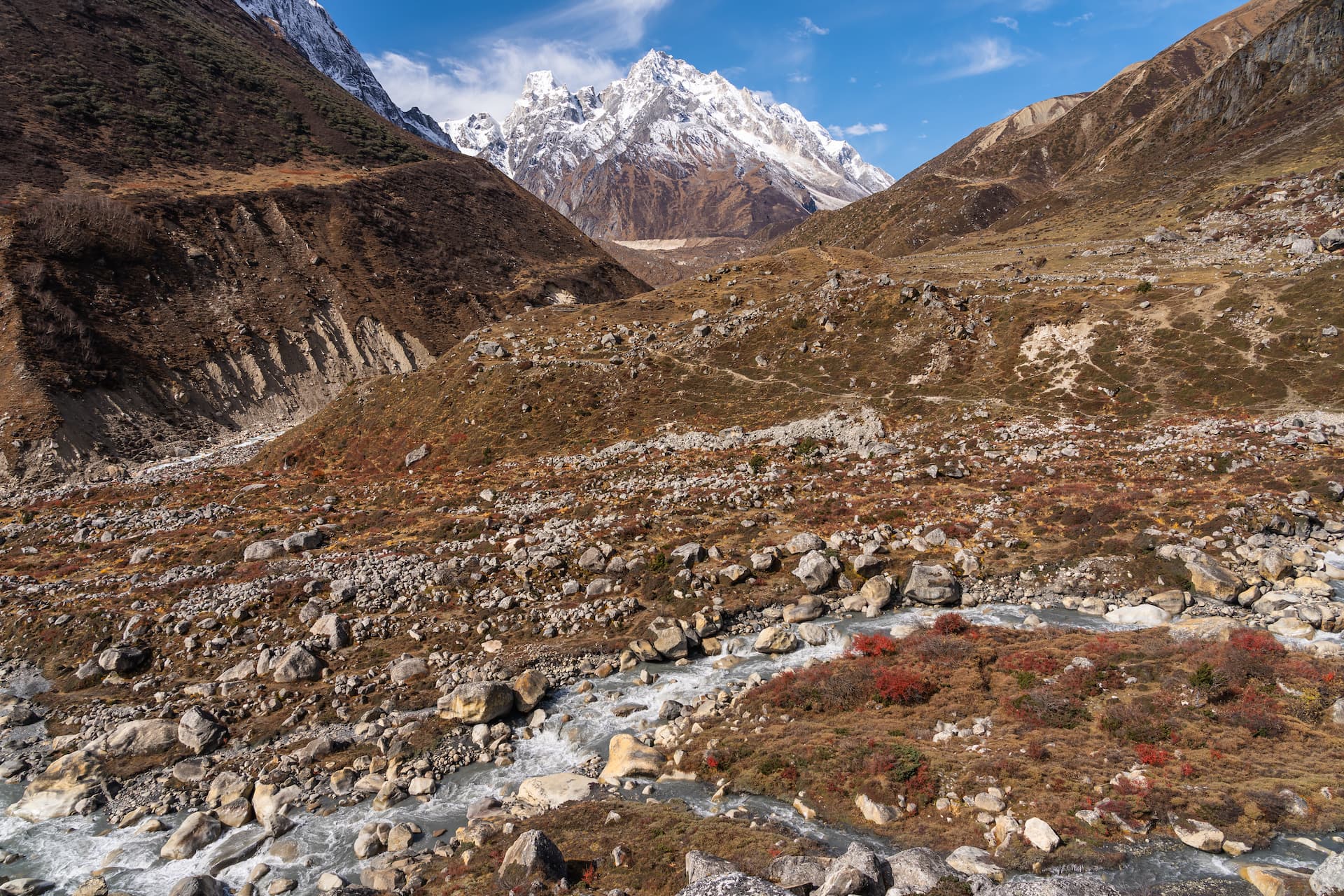 Rocky trekking trail leading toward snow-capped mountains above a rushing river in a high valley