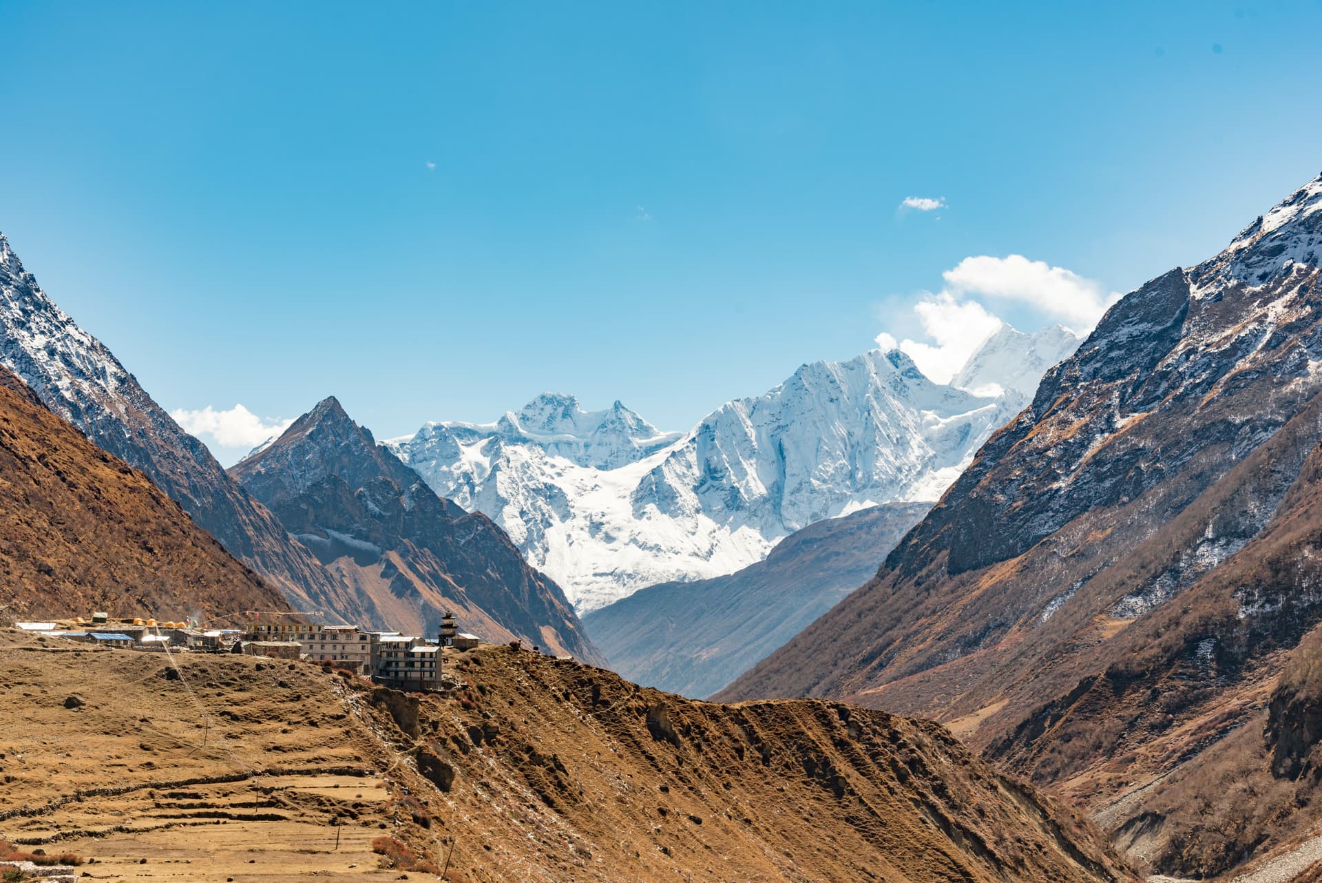 Mountain village nestled in valley below massive snow-covered Himalayan peaks under blue sky.