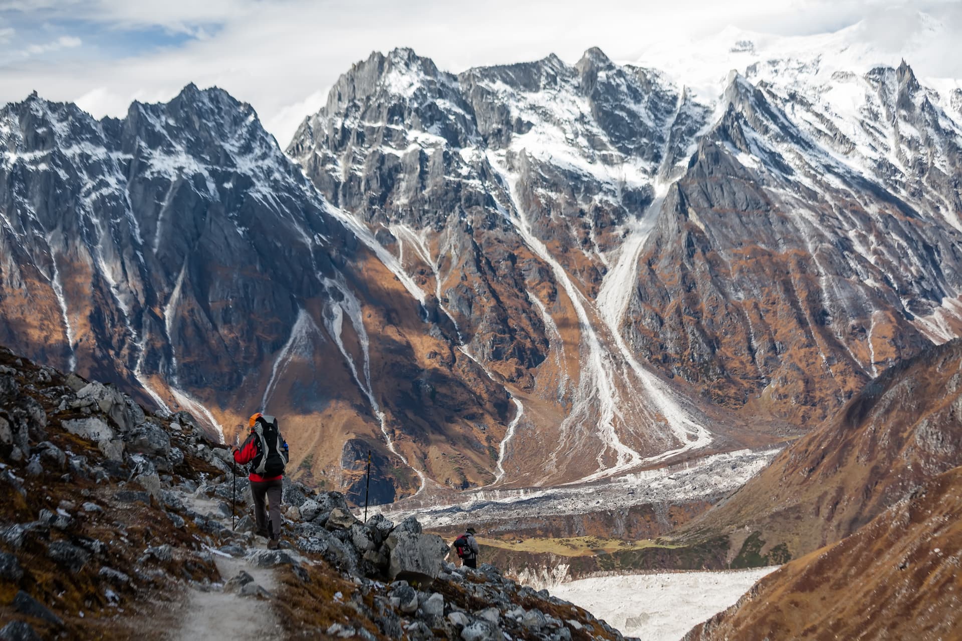 Hikers trekking on rocky trail with massive snow-capped mountains and glacier valley below