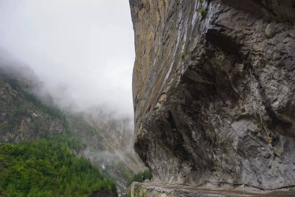 Hikers on narrow road carved into cliff face near Besisahar, shrouded in mountain fog.