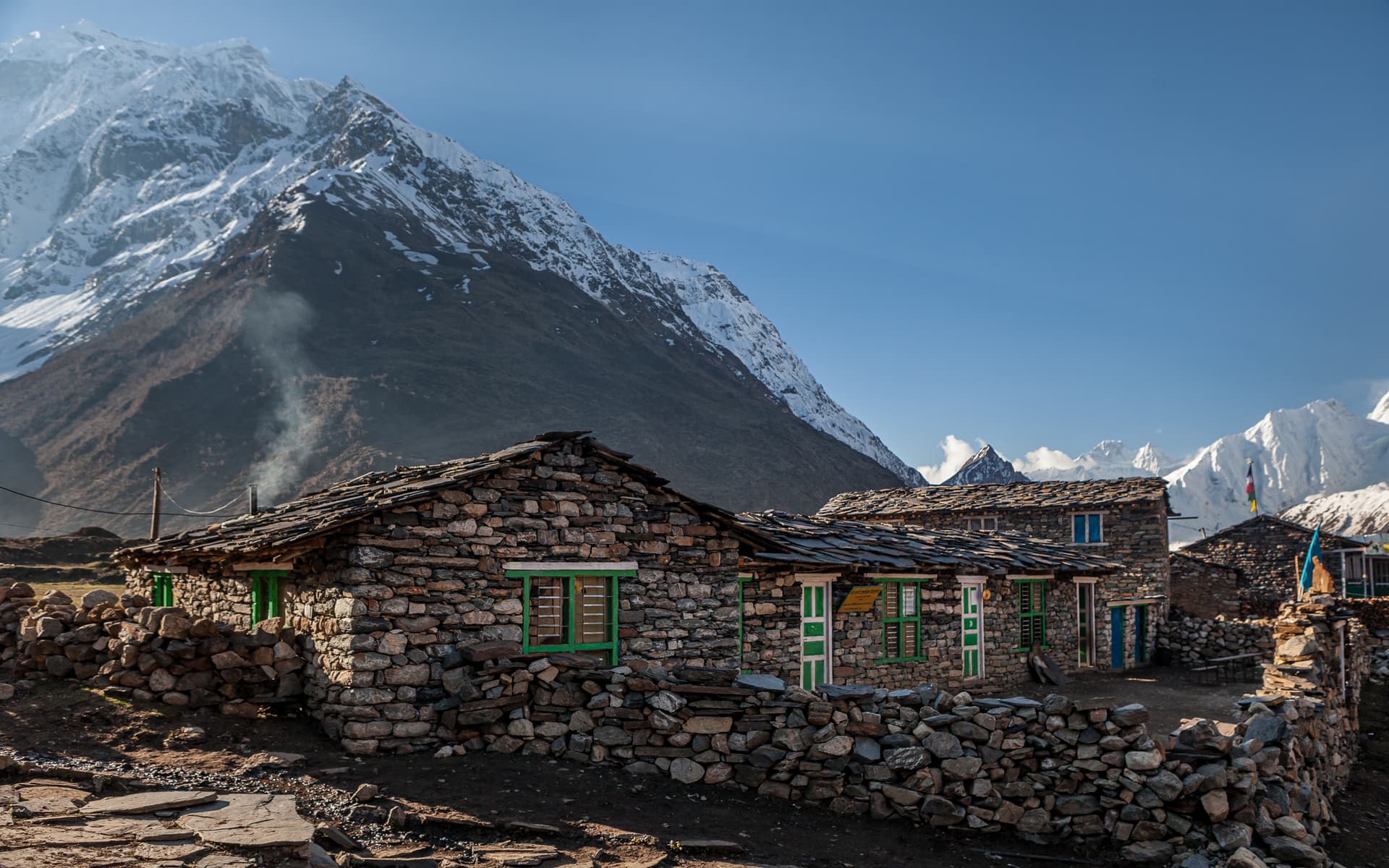 Stone houses with slate roofs in Manaslu region with snow-capped mountains under blue sky.