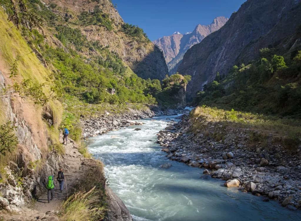 Hikers trekking along a narrow path beside a rushing river in a deep mountain valley with snow-capped peaks.