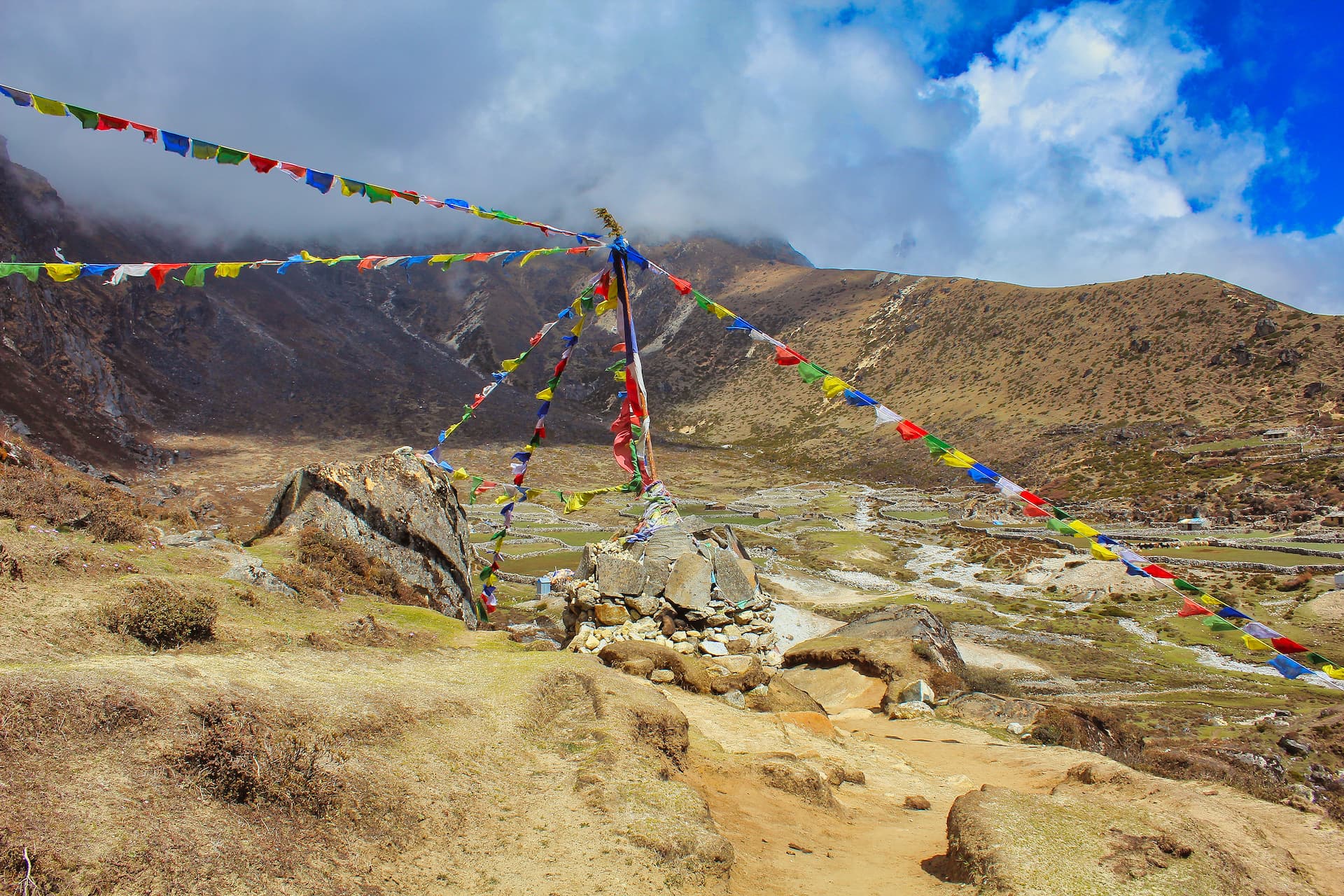 Prayer flags strung across a dry, grassy mountain landscape near Machermo under a cloudy blue sky.
