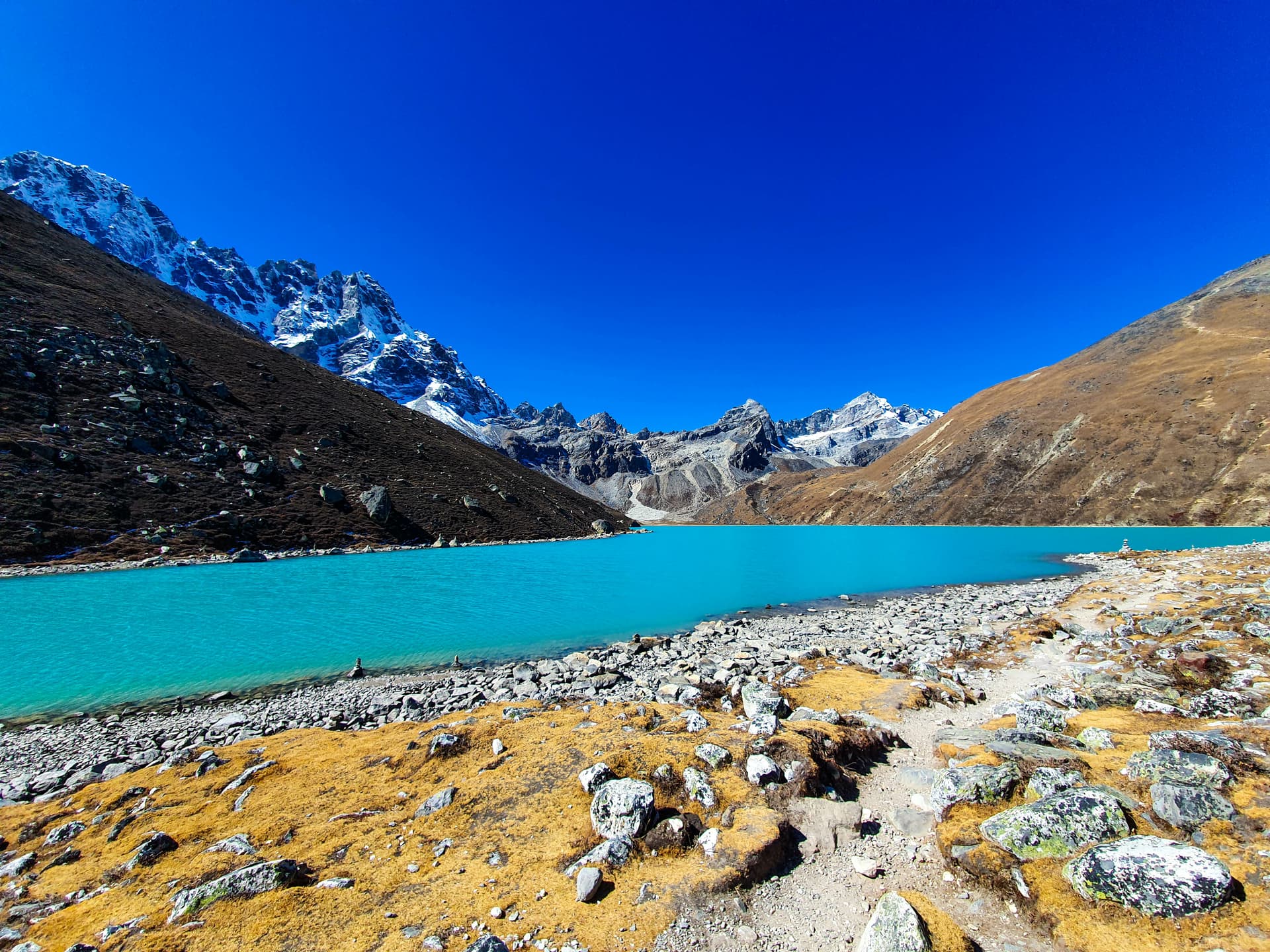 Turquoise alpine lake from Machhermo to Gokyo with snow-capped mountains under bright blue sky.