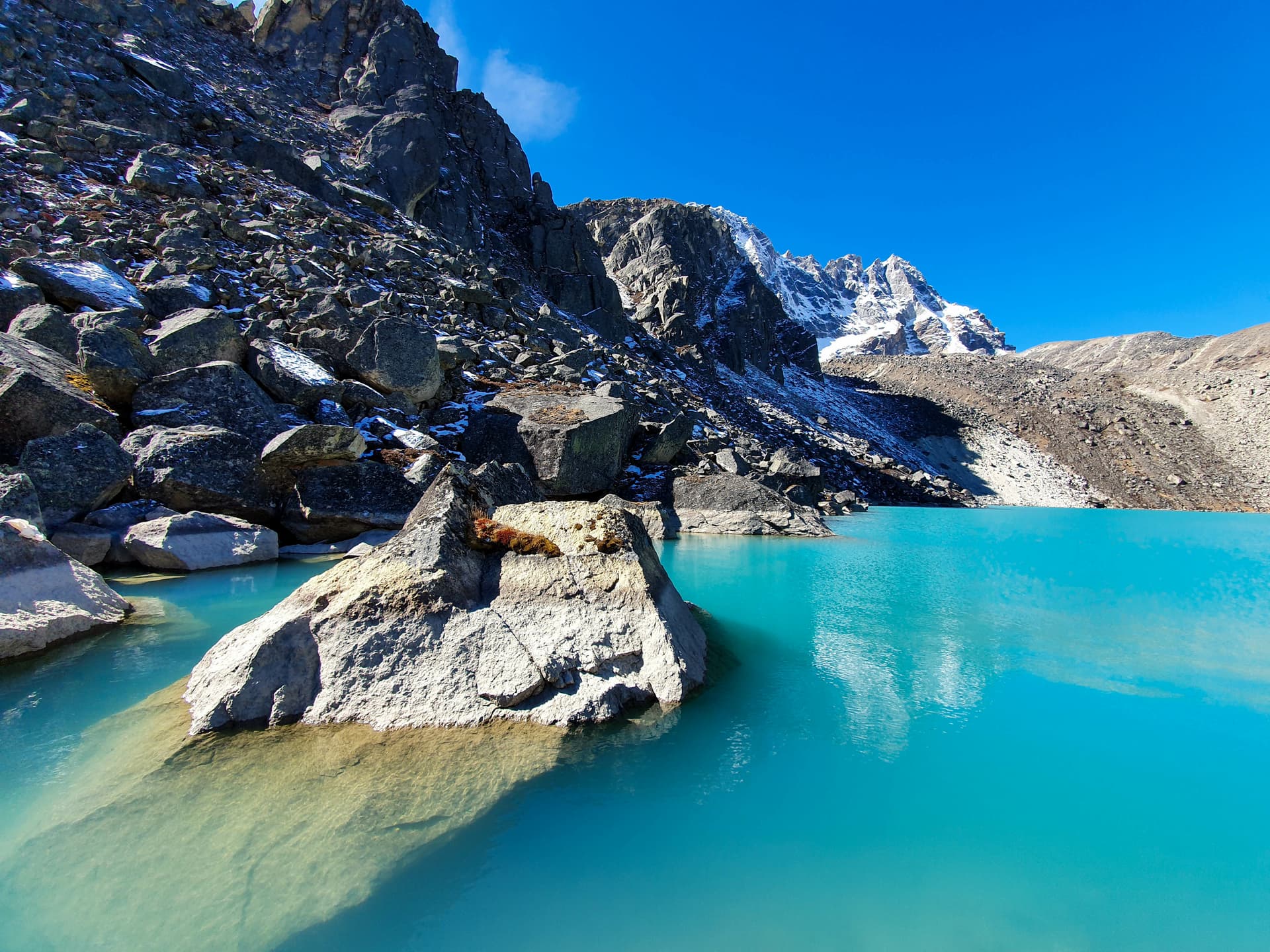 Turquoise Gokyo Lake with rocky shore and snow-dusted mountains under bright blue sky.