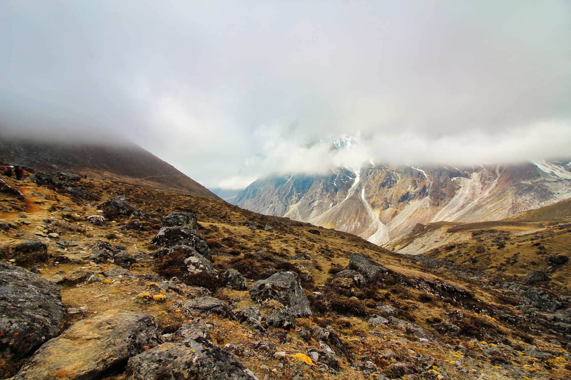 Hikers ascending rocky, grassy slope towards snow-capped mountains shrouded in clouds near Dzonghla.