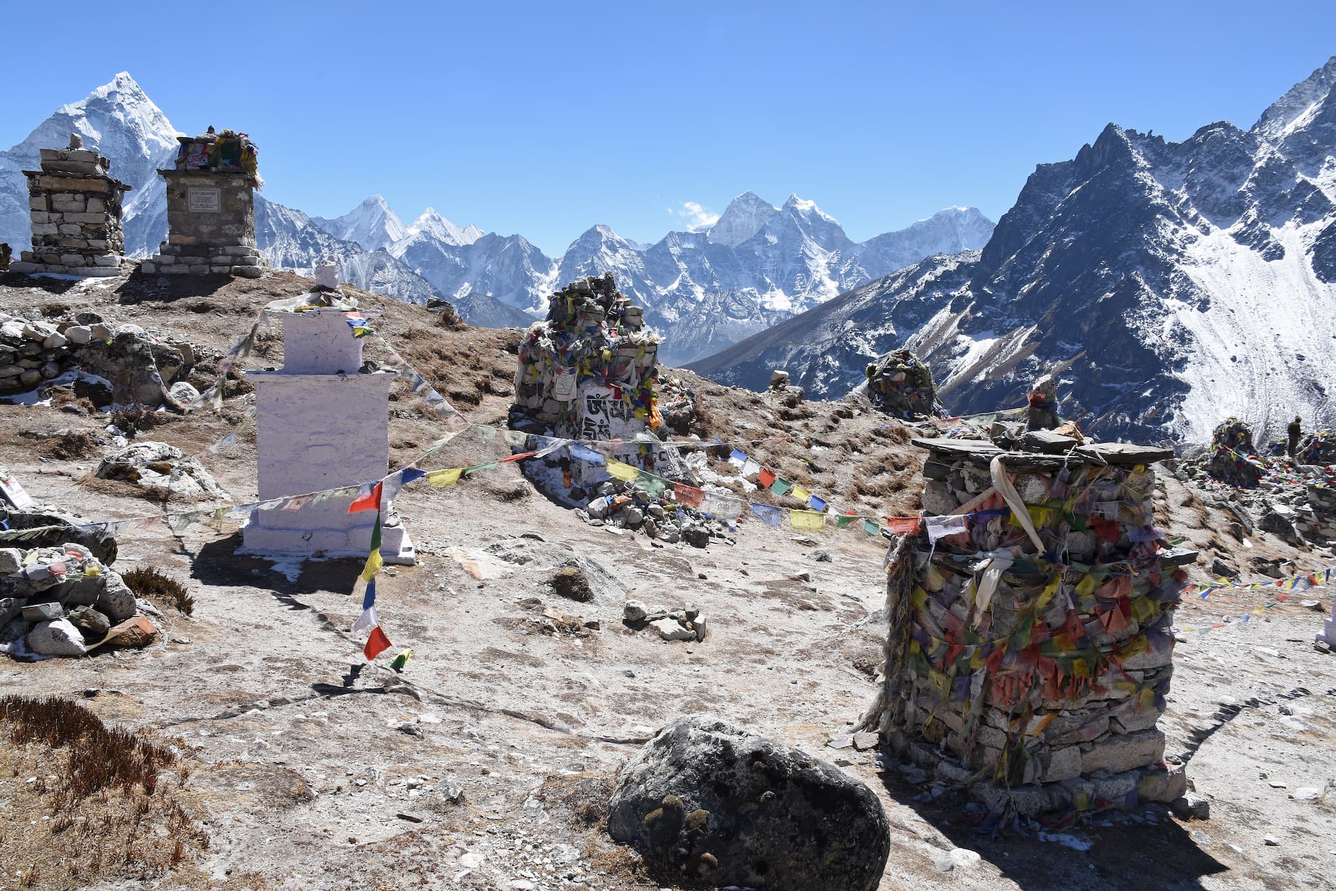 Chortens and prayer flags on barren hillside with snow-capped Himalayan mountains in background.