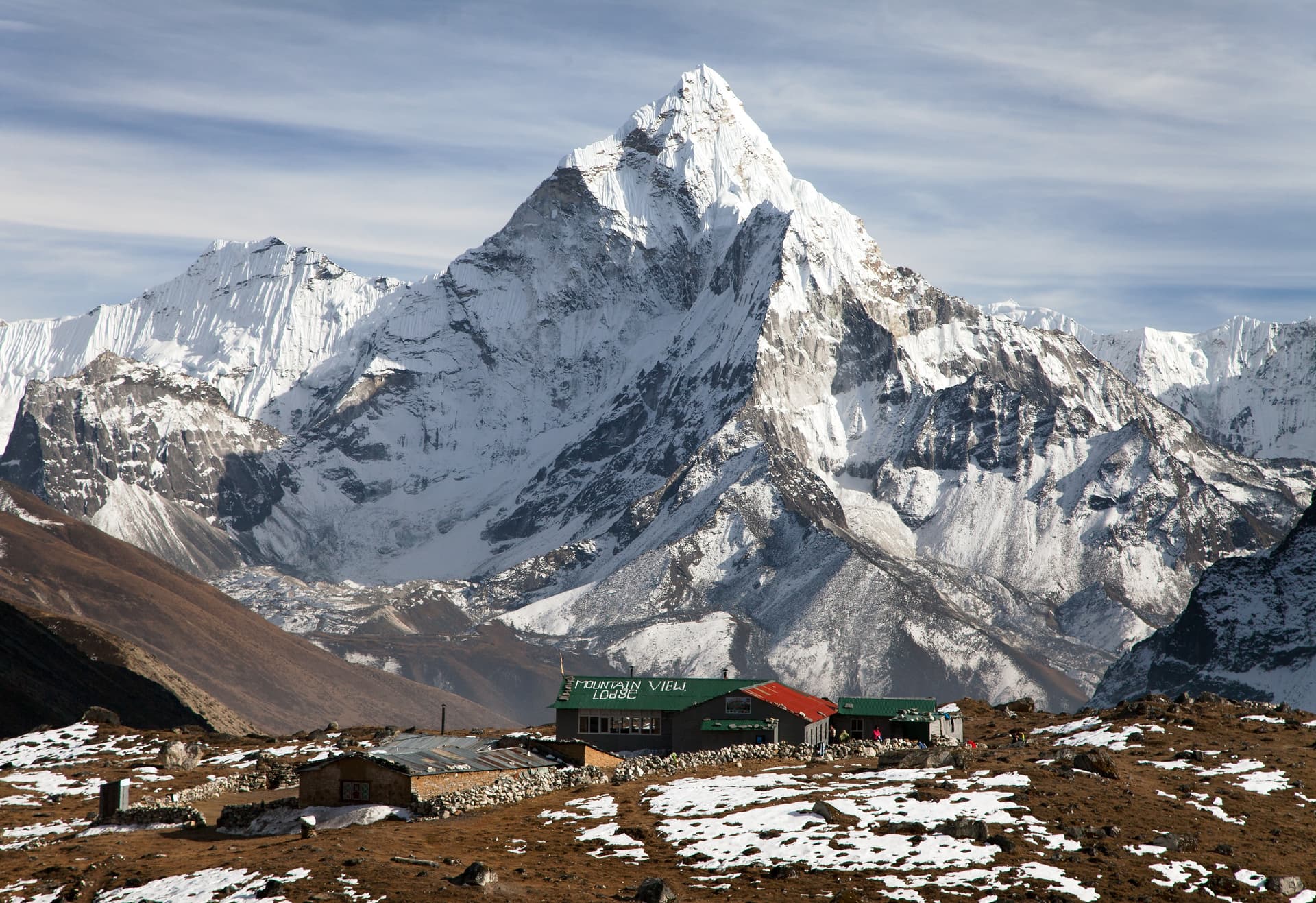 Snow-covered Ama Dablam mountain towering over Mountain View Lodge on brown, patchy snow terrain.